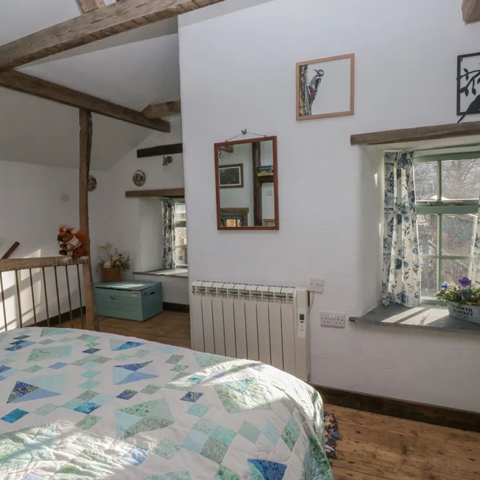 A bedroom with a bed a window with floral curtains a mirror on the wall and wooden beams at The Old Cart House in Garsdale near Sedbergh