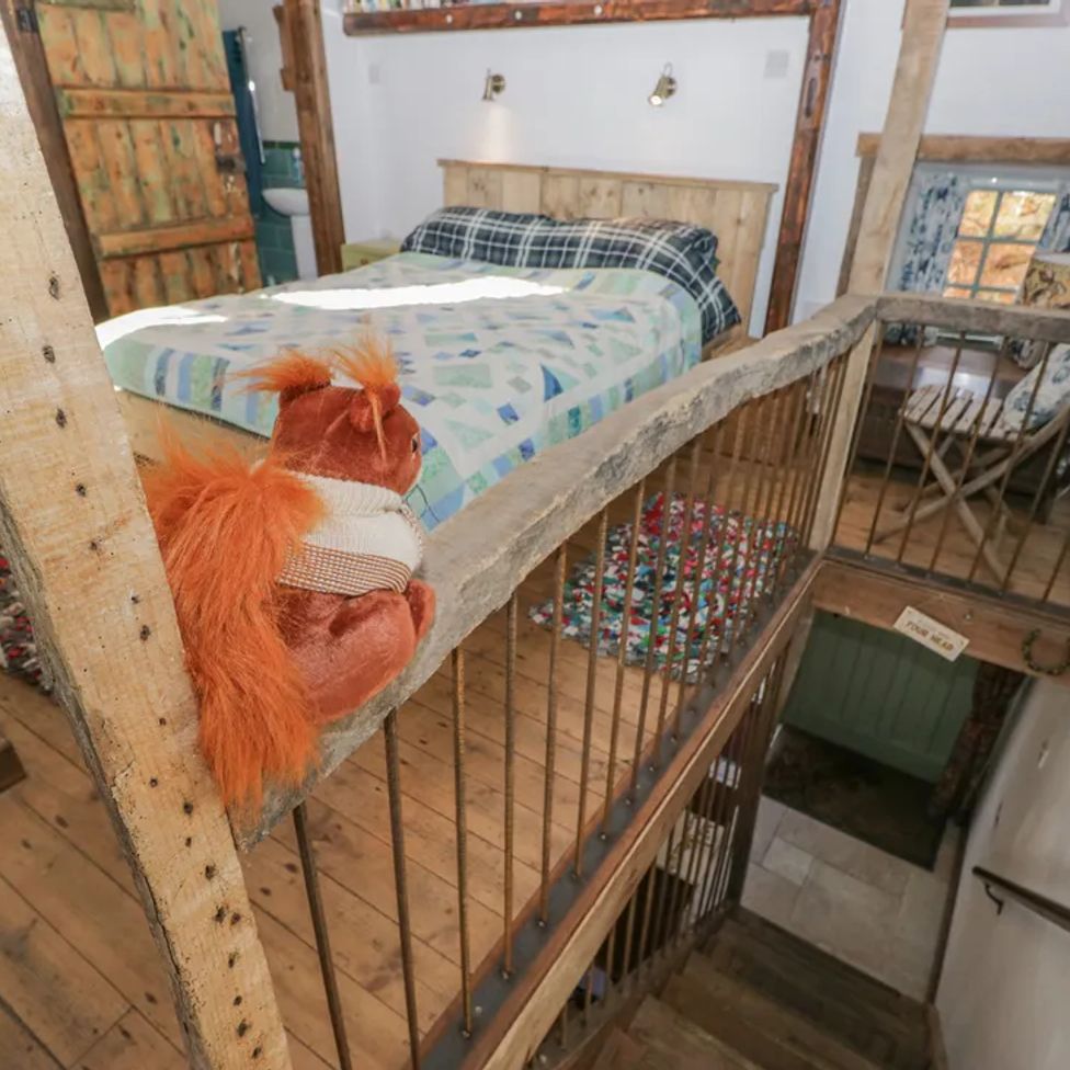 A bedroom with a bed covered in a patterned quilt a toy squirrel on the wooden railing and stairs leading down at The Old Cart House in Garsdale near Sedbergh