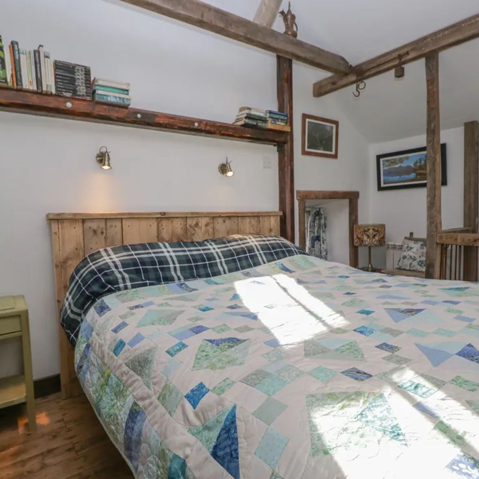 A bedroom with a wooden bed frame quilted bedspread green bedside table books on a wall shelf and framed pictures at The Old Cart House in Garsdale near Sedbergh