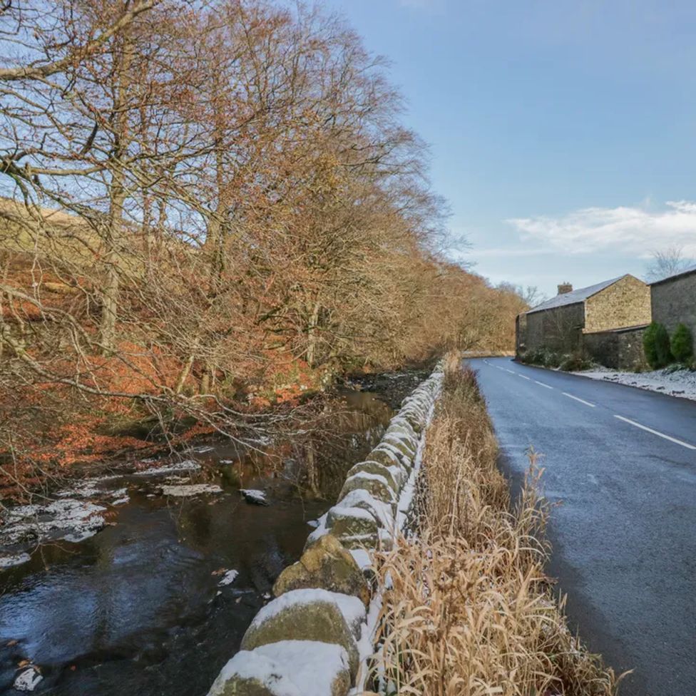 A narrow road alongside a stone wall with a stream and leafless trees on the left and stone buildings on the right at The Old Cart House in Garsdale near Sedbergh
