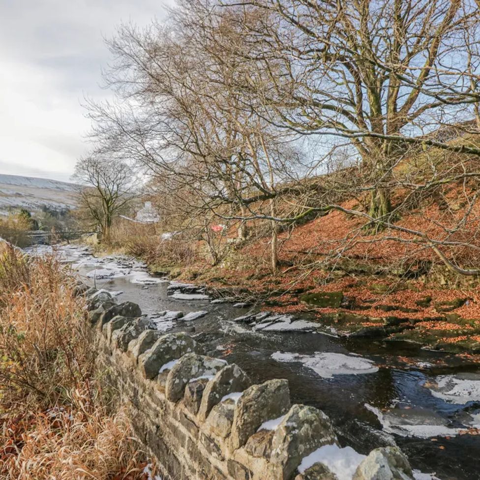 A road running alongside a stone wall and a partially frozen stream with leafless trees at The Old Cart House in Garsdale near Sedbergh