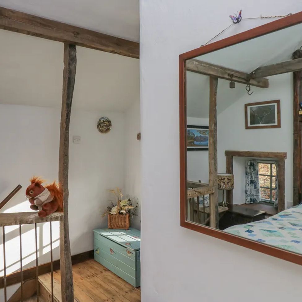 A staircase with wooden railing and a stuffed squirrel on the banister by a green chest with flowers in a basket and a mirror reflecting a bedroom with wooden beams at The Old Cart House in Garsdale near Sedbergh