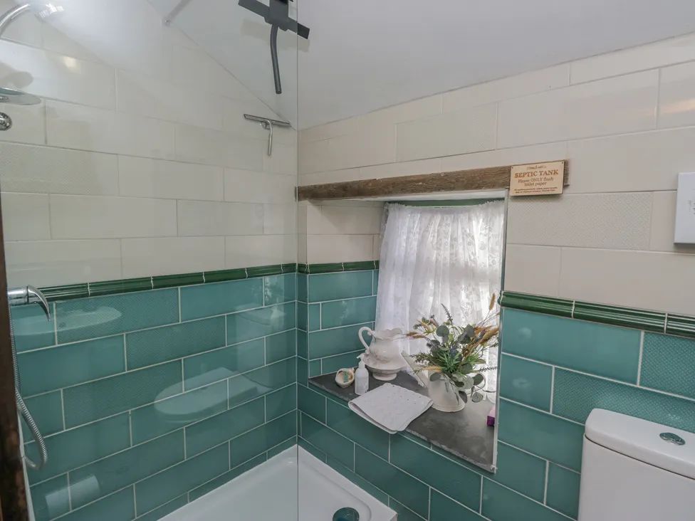 A bathroom with a shower area green and white tiled walls a small window with a curtain flowers and toiletries on the window sill and a toilet at The Old Cart House in Garsdale near Sedbergh