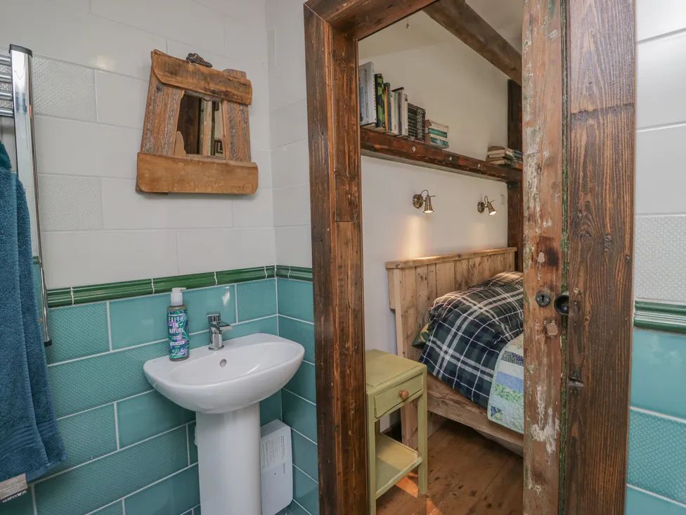 A bathroom with a sink and mirror next to a wooden door opening to a bedroom with a bed and bookshelf at The Old Cart House in Garsdale near Sedbergh
