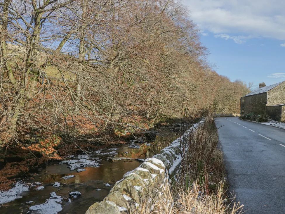 A rural road with a stone wall beside a stream and leafless trees near a stone building at The Old Cart House in Garsdale near Sedbergh