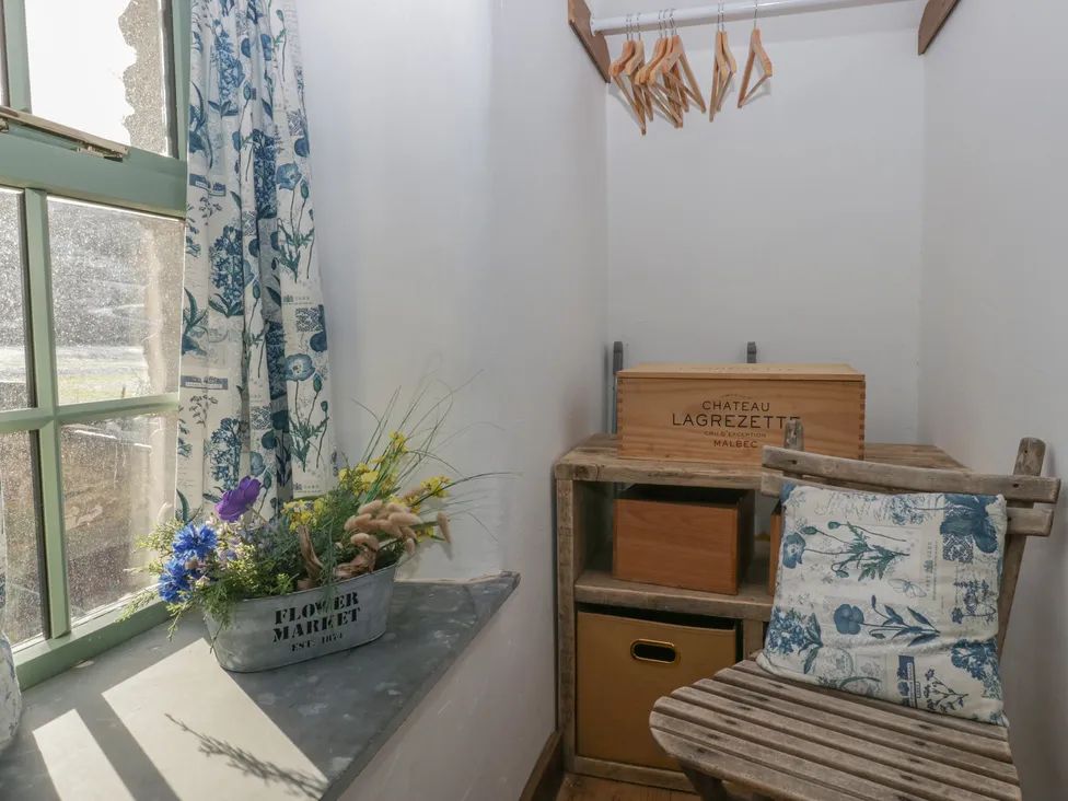 A small corner with a window, a flowerpot on the sill, a wooden chair with a cushion, hangers, and storage boxes at The Old Cart House in Garsdale near Sedbergh