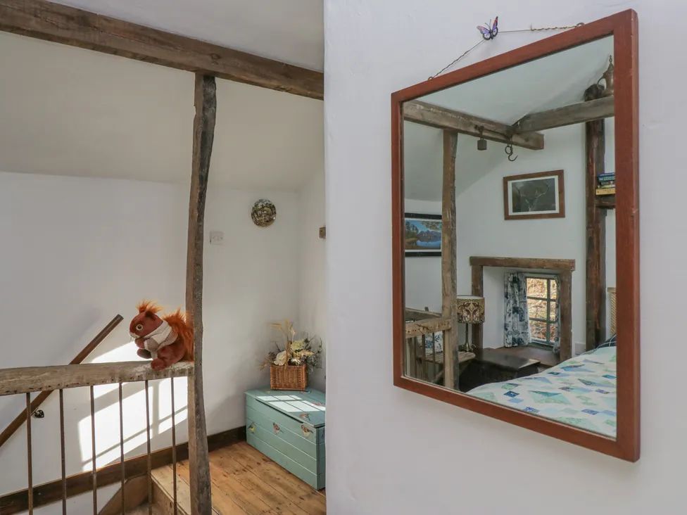 A staircase with wooden railing and a stuffed squirrel on the banister by a green chest with flowers in a basket and a mirror reflecting a bedroom with wooden beams at The Old Cart House in Garsdale near Sedbergh